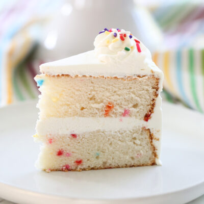 A piece of funfetti cake with white frosting and sprinkles on a white plate.